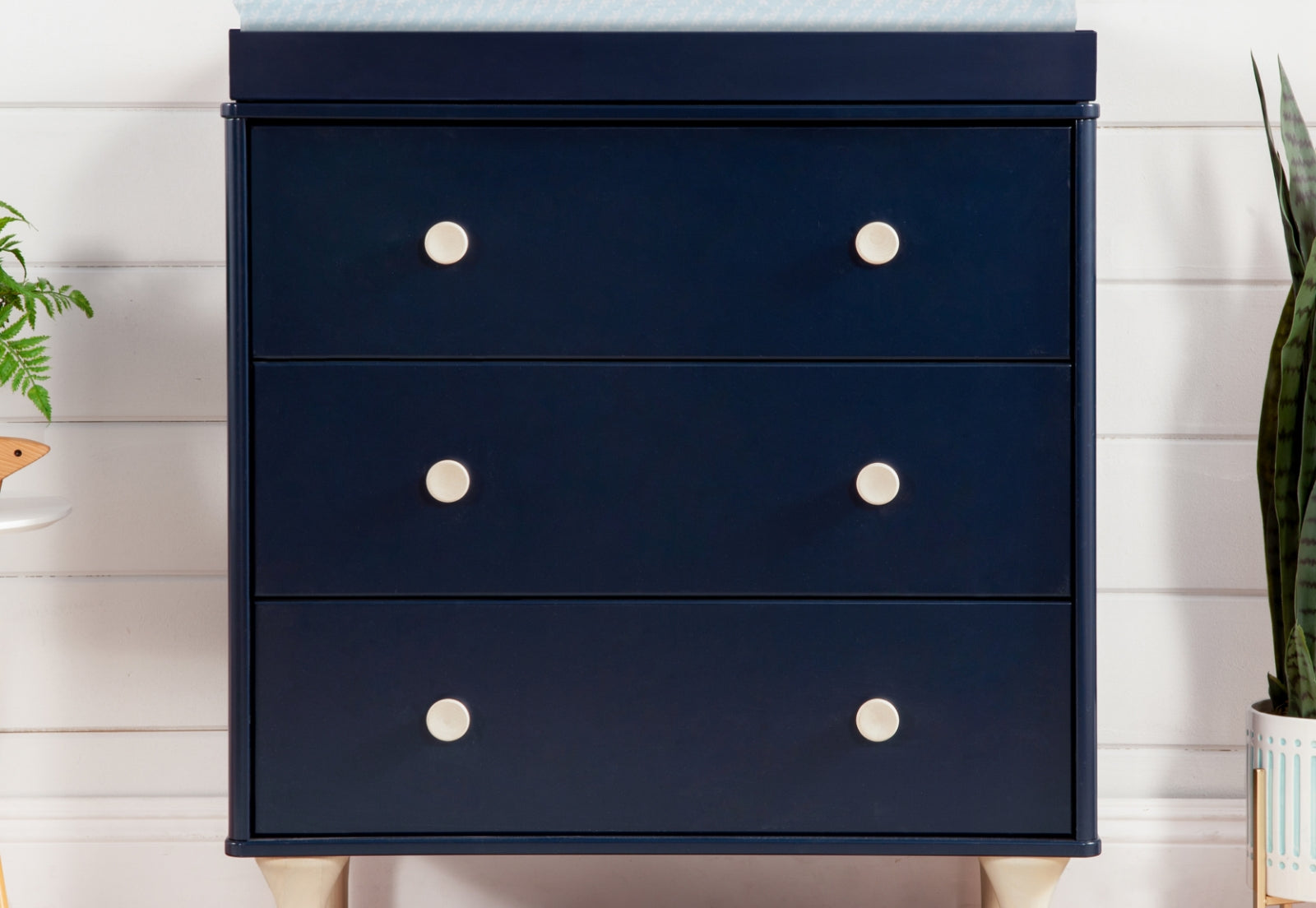 A navy blue modern dresser with three drawers, each featuring a white circular knob, set against a white paneled wall with a decorative plant to the side.