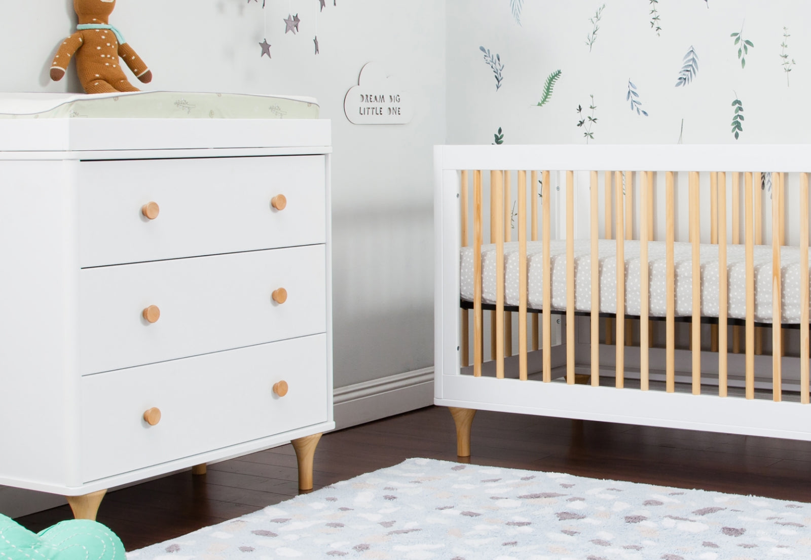 A stylish and serene nursery featuring a white dresser with wooden knobs and a white crib with natural wood spindles, plus a wall decoration that reads "Dream Big Little One."