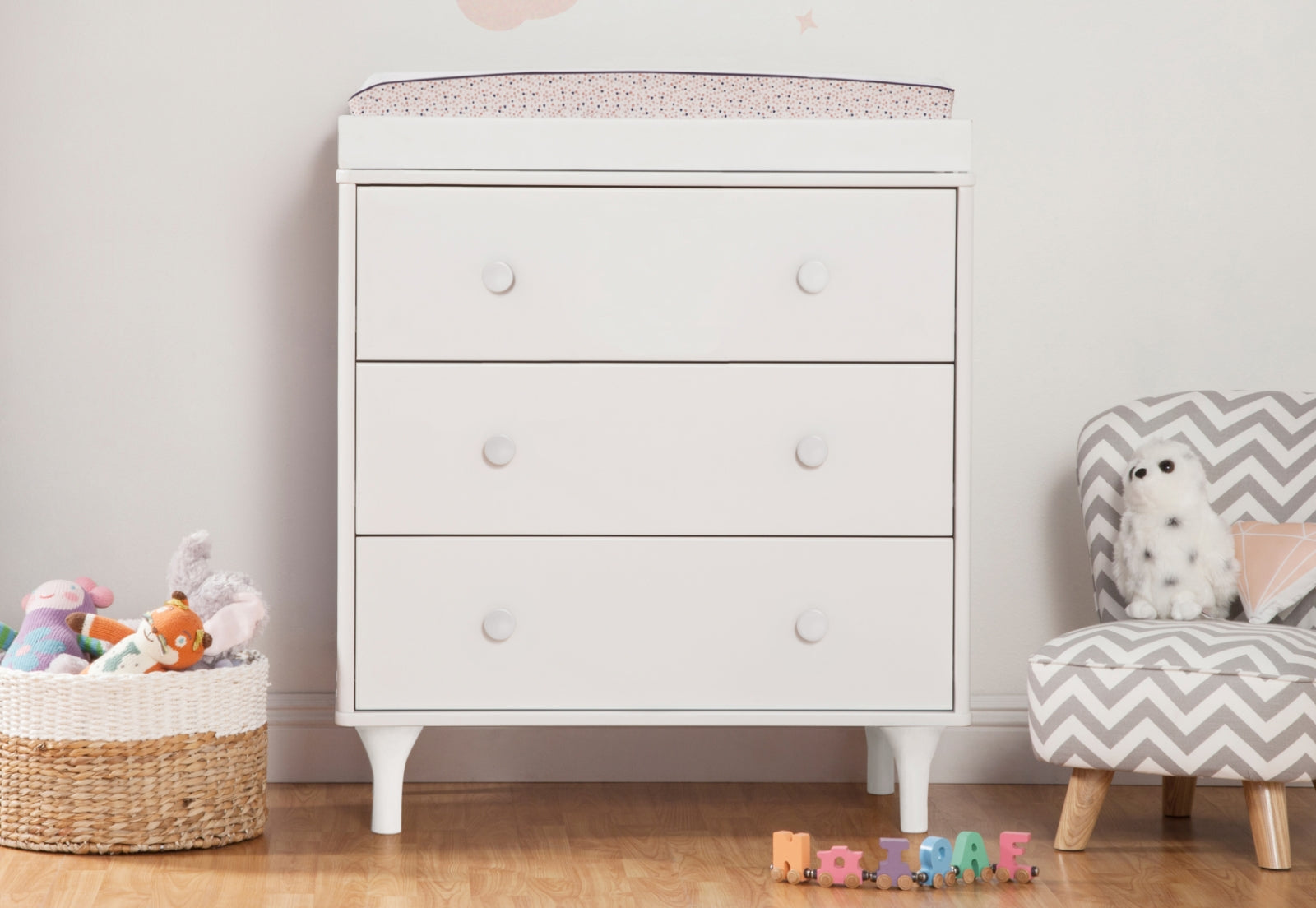 White baby changing table with four drawers in a nursery, accompanied by a basket of toys, a chevron-patterned chair with a stuffed toy, and colorful wooden blocks spelling out "NATHAN."