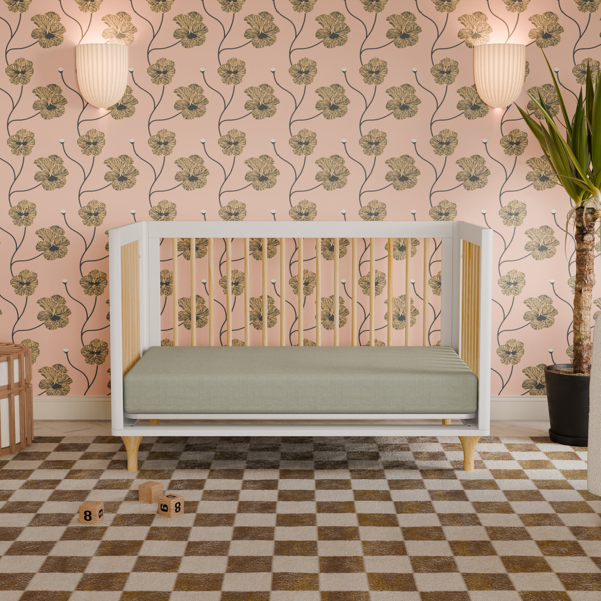 The Babyletto Lolly Convertible Crib, crafted from sustainable New Zealand pine wood in a modern white finish, is featured in a nursery with a green mattress, floral wallpaper, wall sconces, a corner plant, checkered rug, and wooden blocks scattered on the floor.