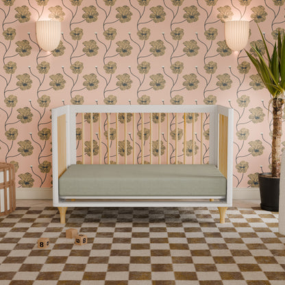The Babyletto Lolly Convertible Crib, crafted from sustainable New Zealand pine wood in a modern white finish, is featured in a nursery with a green mattress, floral wallpaper, wall sconces, a corner plant, checkered rug, and wooden blocks scattered on the floor.