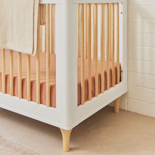 The Babyletto Lolly Convertible Crib, crafted from New Zealand pine wood, features light wood legs and bars and sits in a softly lit room with beige walls. Fitted with a peach-colored sheet and adorned with a tan knit blanket draped over the side, this elegant crib beautifully blends white and natural wood tones, creating a serene nursery atmosphere.