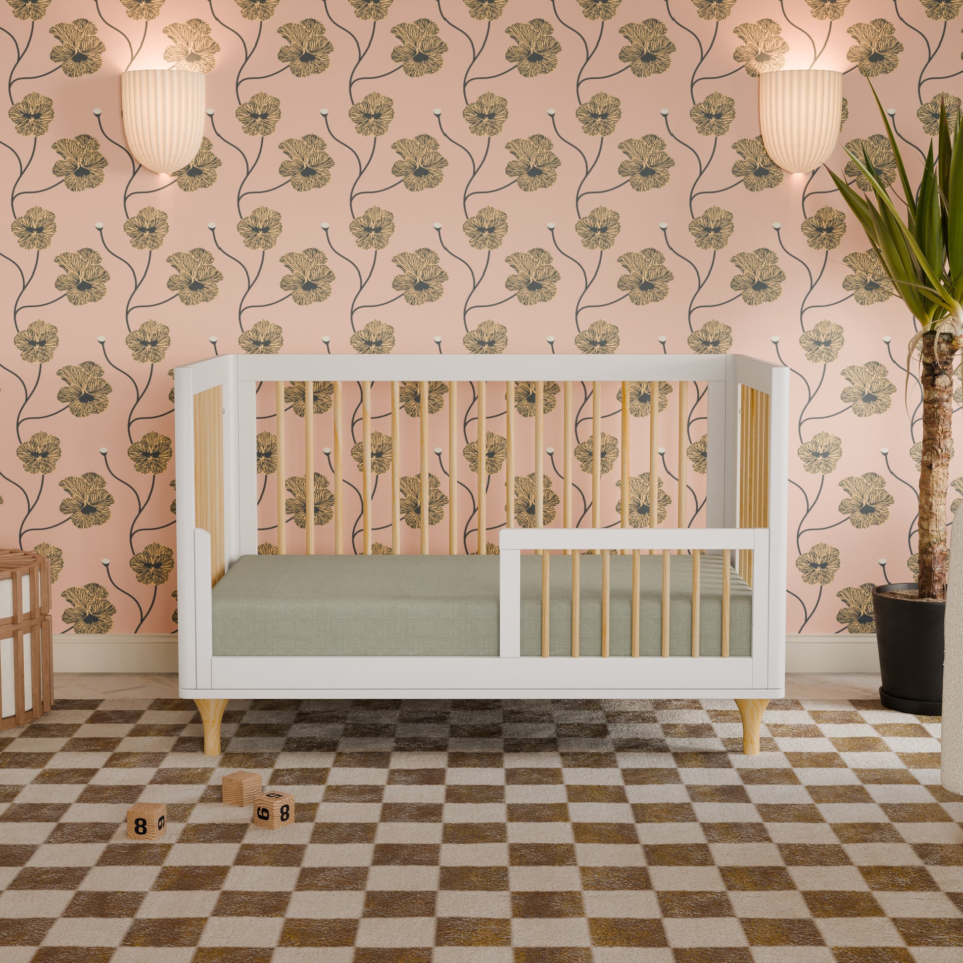 A sleek white and natural wood Lolly Convertible Crib by Babyletto, crafted from sustainable New Zealand pine, sits on a checkered rug against pink floral wallpaper. Two wall lights and a tall potted plant are in the background, with toy blocks scattered on the floor in front of the crib.
