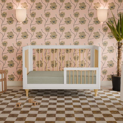 A sleek white and natural wood Lolly Convertible Crib by Babyletto, crafted from sustainable New Zealand pine, sits on a checkered rug against pink floral wallpaper. Two wall lights and a tall potted plant are in the background, with toy blocks scattered on the floor in front of the crib.