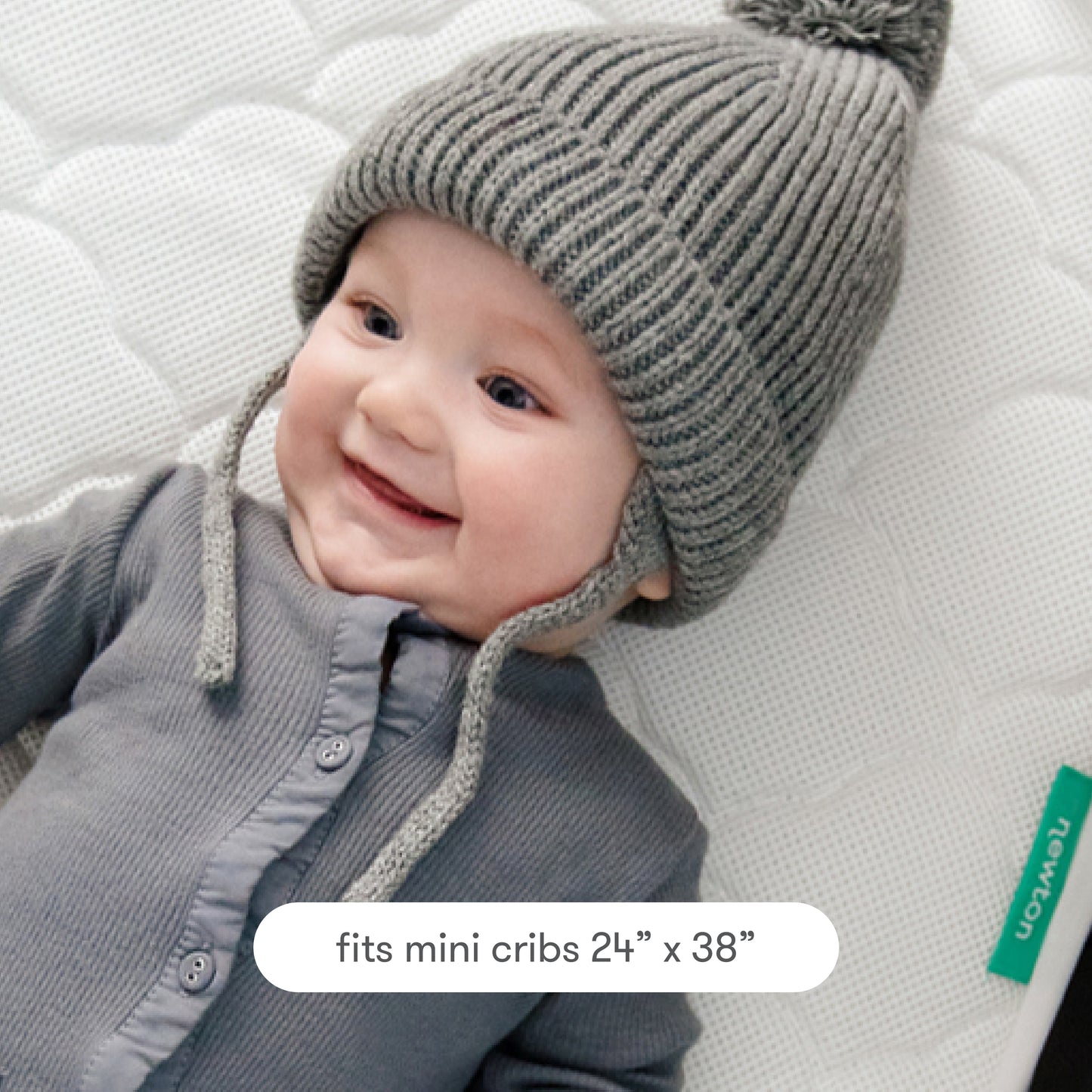 baby in grey clothing and grey beanie hat smiling and lying on newton mini crib mattress pad, with text "fits mini cribs 24" x 38""