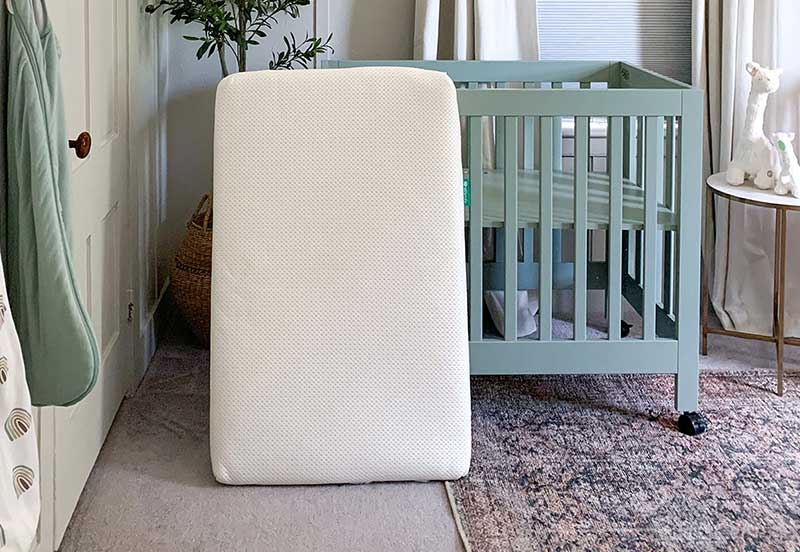 A breathable crib mattress from Newton Baby stands upright in a nursery room near a teal crib, styled with a fittingly serene and nurturing décor.