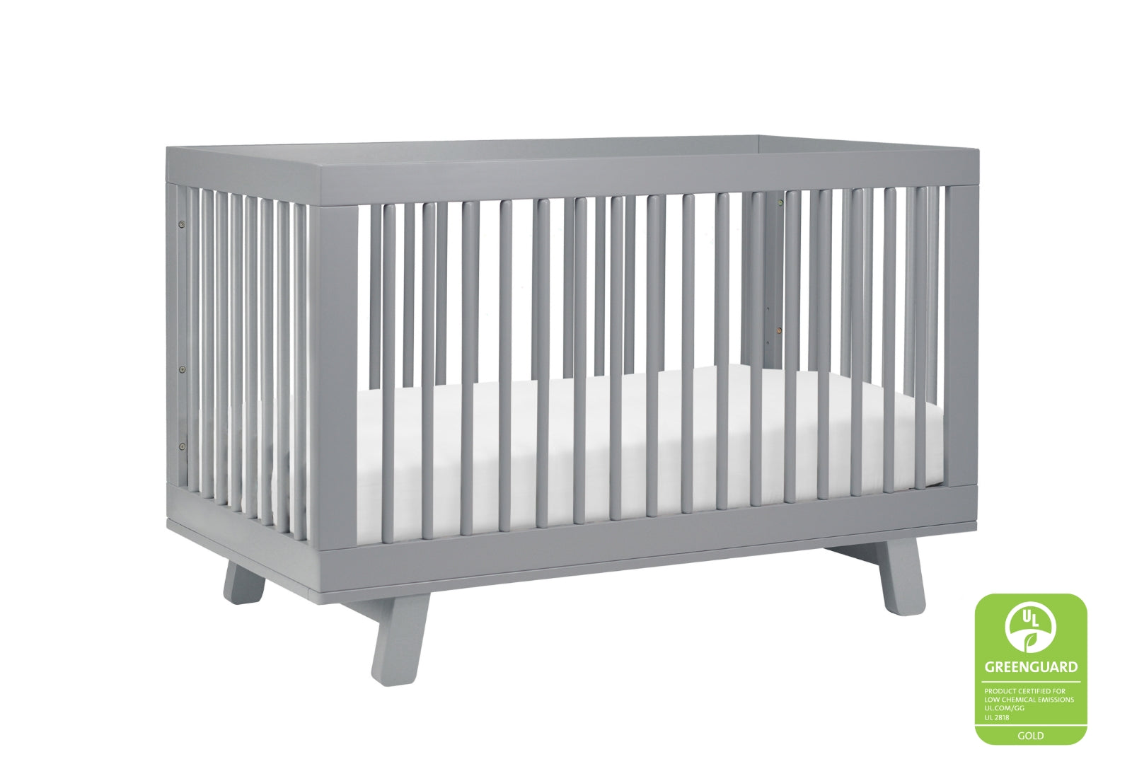 A modern gray crib with a white mattress, featuring vertical bars and a GREENGUARD Gold certification logo for low chemical emissions.