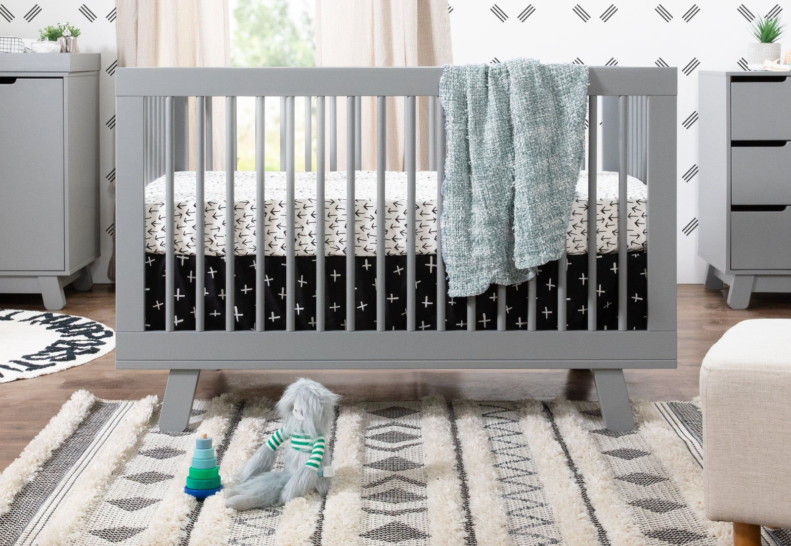 A stylish baby nursery featuring a modern gray crib with star-patterned bedding, a teal blanket, toys on a geometric rug, and neutral-toned furniture.
