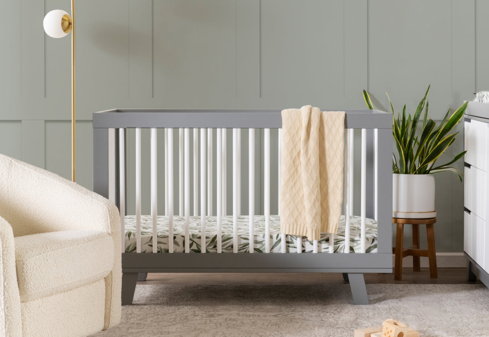 Gray baby crib with a beige blanket hanging on it, located in a nursery room with sage green walls, a white armchair, and a potted plant. 999-8501-GYW