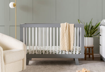 Gray baby crib with a beige blanket hanging on it, located in a nursery room with sage green walls, a white armchair, and a potted plant. 999-8501-GYW