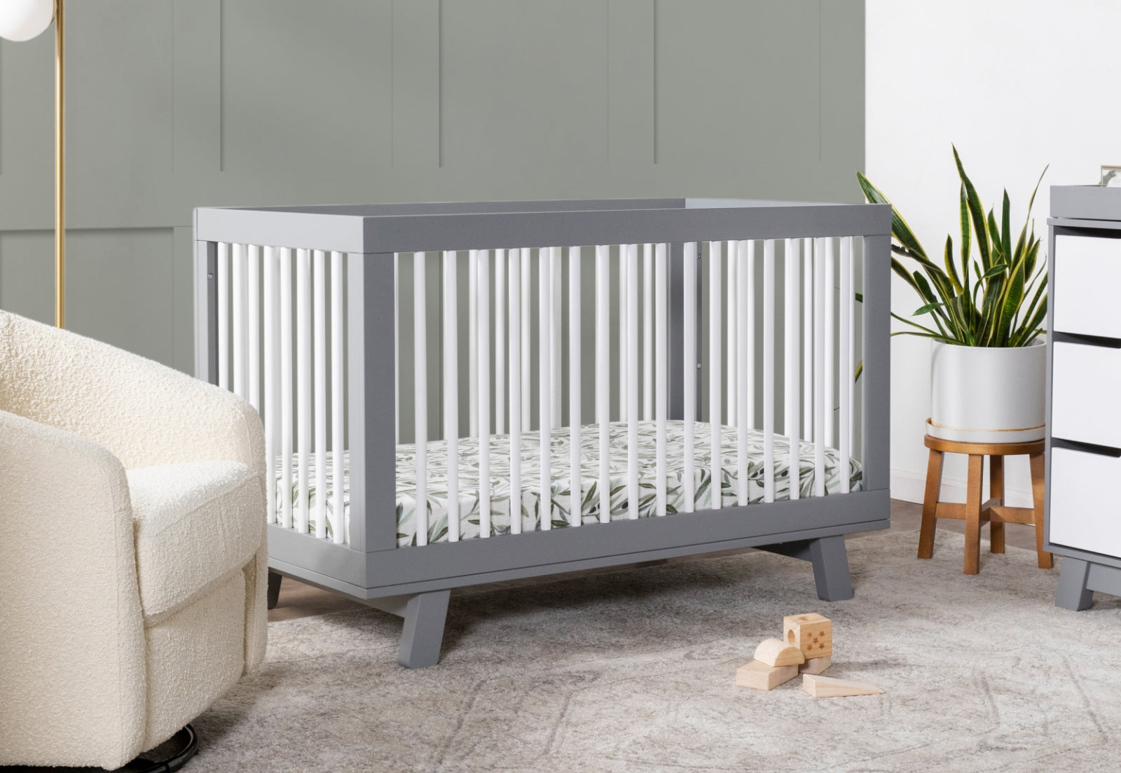 A modern nursery featuring a gray crib with white railings, surrounded by a plush chair, houseplant, and simple toys on a textured rug.