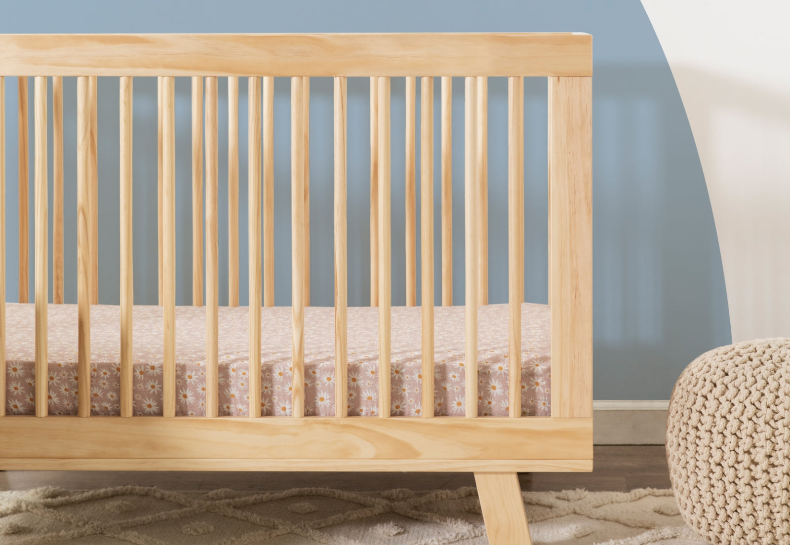 A wooden baby crib with a floral patterned mattress set in a serene room with a beige area rug and knitted pouf.