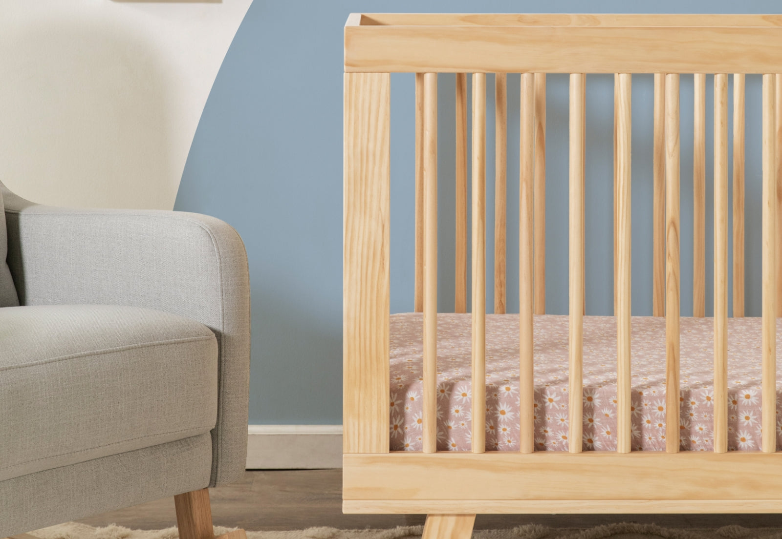 Wooden crib with visible grain beside a modern gray armchair, set against a two-tone wall of cream and blue.