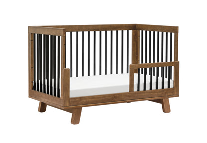 Modern wooden baby crib with visible slats and a lowered side gate on a white background.