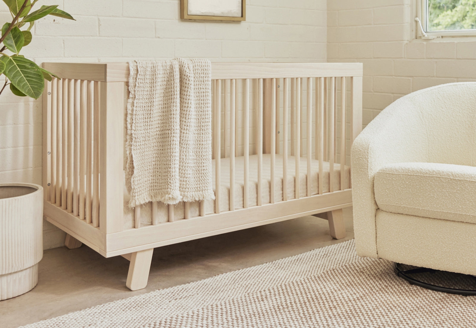 A cozy nursery featuring a modern wooden crib draped with a knit blanket, next to a comfortable armchair and under a bright window. 999-8501-WNA