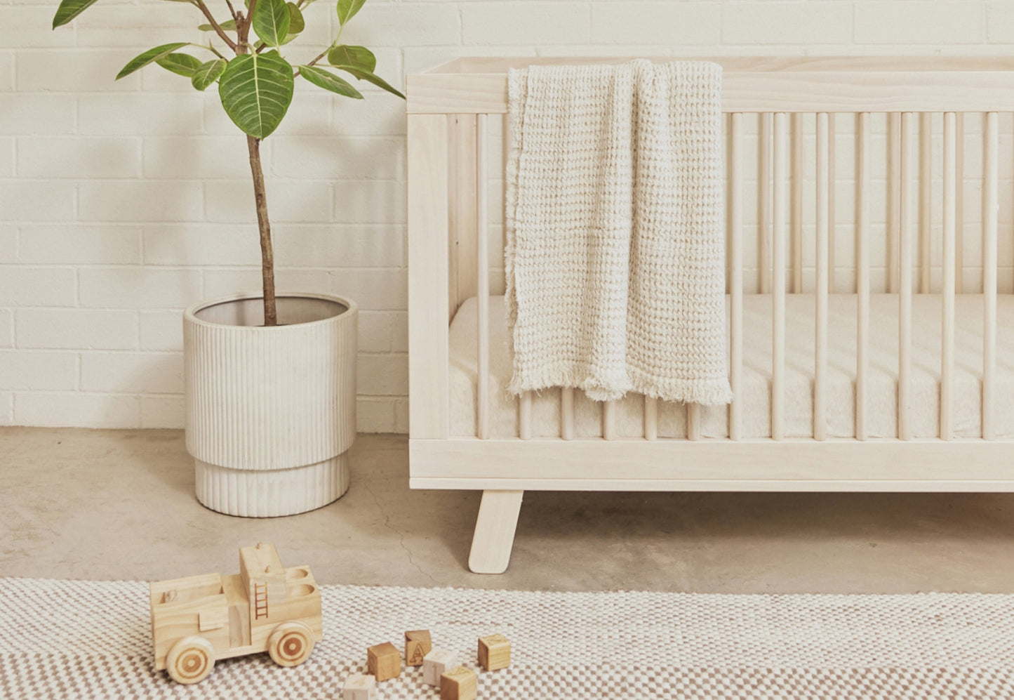 A cozy nursery scene featuring a wooden crib draped with a knitted blanket, a plant in a white pot, wooden toys, and blocks on a textured rug, set against a white brick wall.