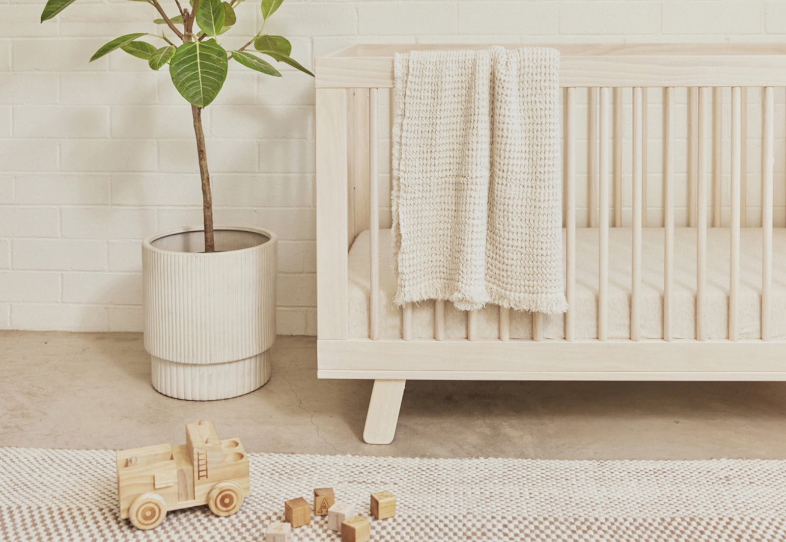 A cozy nursery scene featuring a wooden crib draped with a knitted blanket, a plant in a white pot, wooden toys, and blocks on a textured rug, set against a white brick wall.