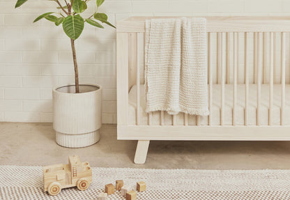 A cozy nursery scene featuring a wooden crib draped with a knitted blanket, a plant in a white pot, wooden toys, and blocks on a textured rug, set against a white brick wall.
