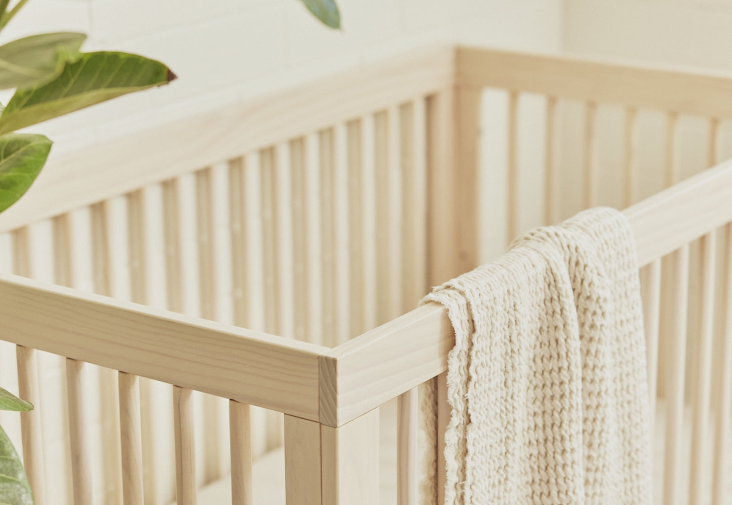 A cream-colored knit blanket draped over the side of a wooden baby crib in a sunny room with a hint of green foliage in the background.