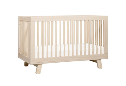 Alt text: "Light wood baby crib with visible grain and angled legs, featuring vertical slats on all sides."
