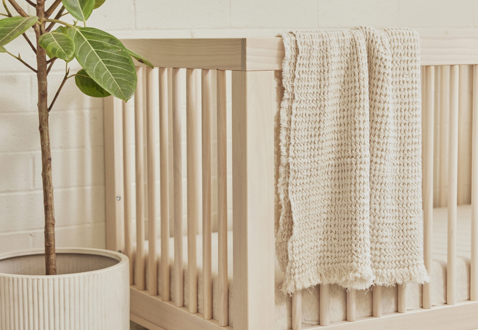 A cozy beige blanket draped over a wooden crib next to a potted fiddle leaf fig plant in a serene nursery setting.