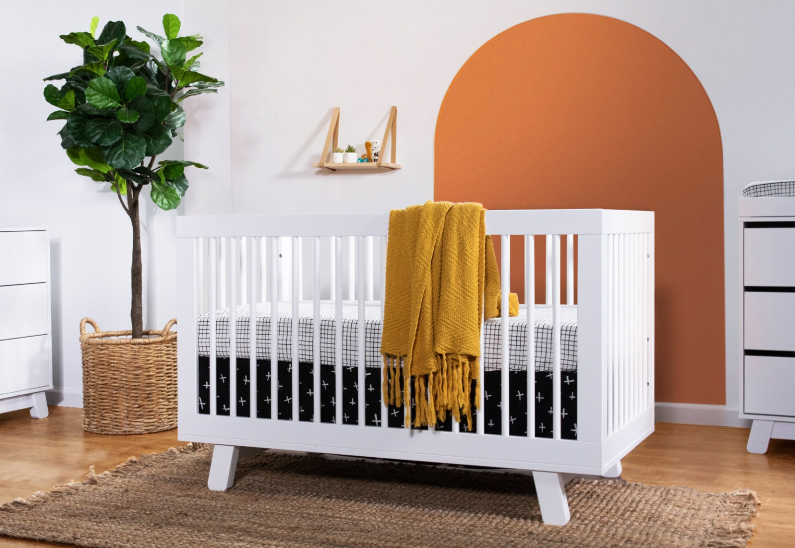Modern nursery with a white crib, decorative mustard yellow throw, and geometric-patterned bedding, accented by a large green plant and stylish orange half-circle wall paint. 999-8501-WHT