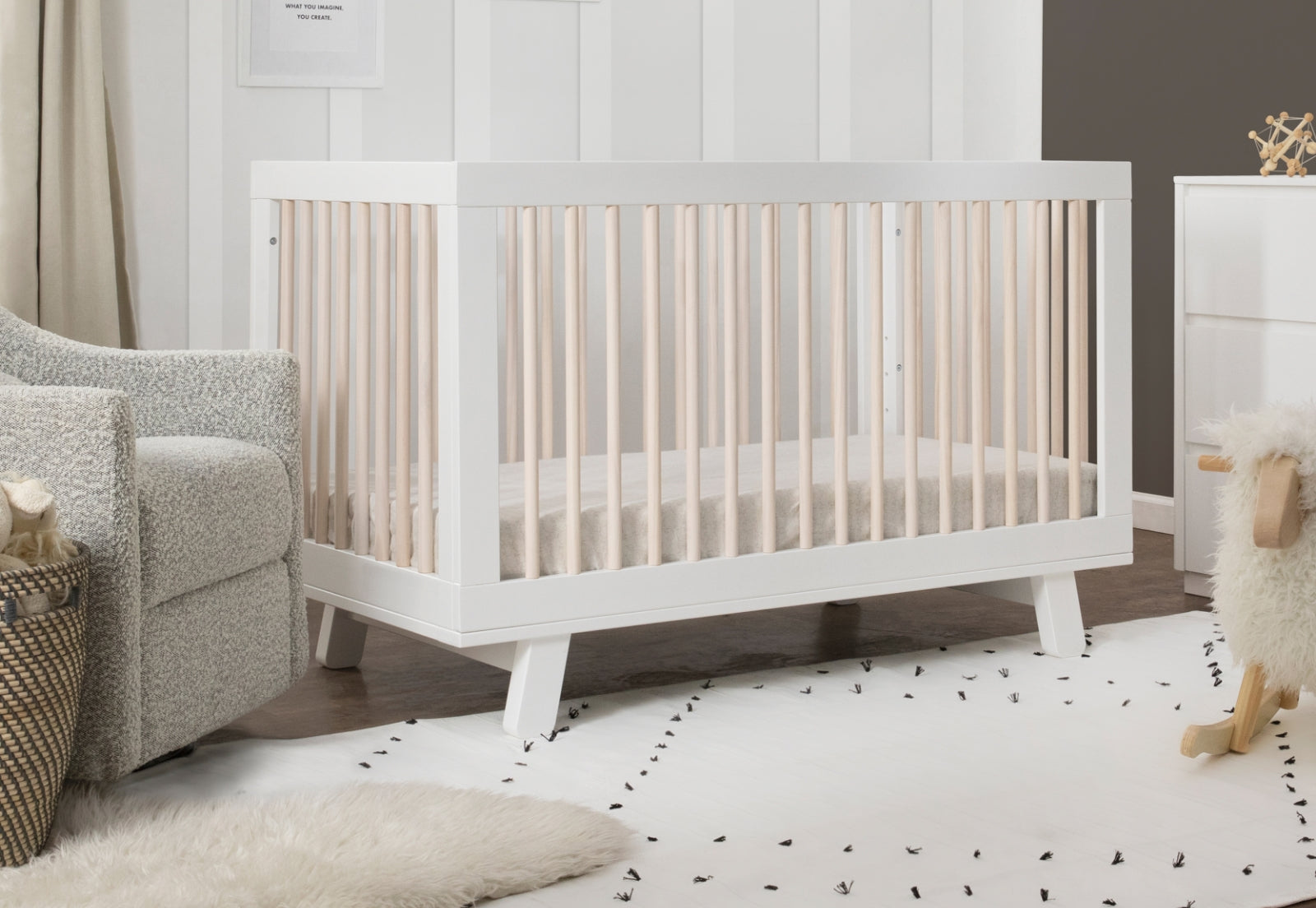 Modern white baby crib in a serene nursery setting with a grey sofa, white dresser, and playful nursery decor. 999-8501-WWN