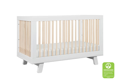 Modern white baby crib with natural wood spindles, featuring a Greenguard Gold certification sticker.