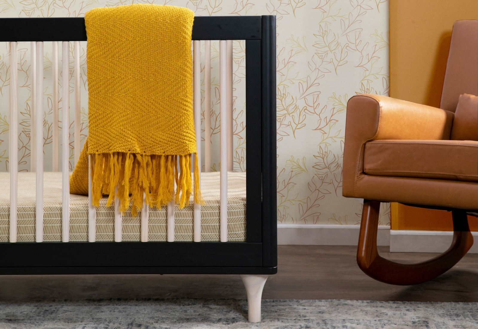 A cozy nursery setup featuring a black crib with a bright yellow throw blanket and a modern brown leather armchair, set against a wallpaper with leaf patterns.