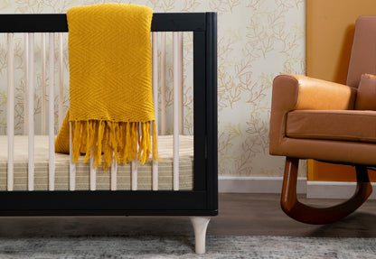 A cozy nursery setup featuring a black crib with a bright yellow throw blanket and a modern brown leather armchair, set against a wallpaper with leaf patterns.