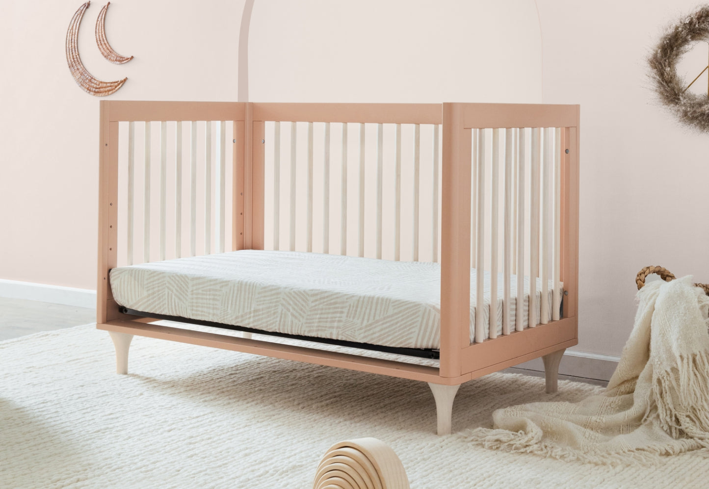 A modern pale pink toddler bed with vertical slats, positioned in a softly decorated room with moon-shaped wall hangings and a plush white rug.