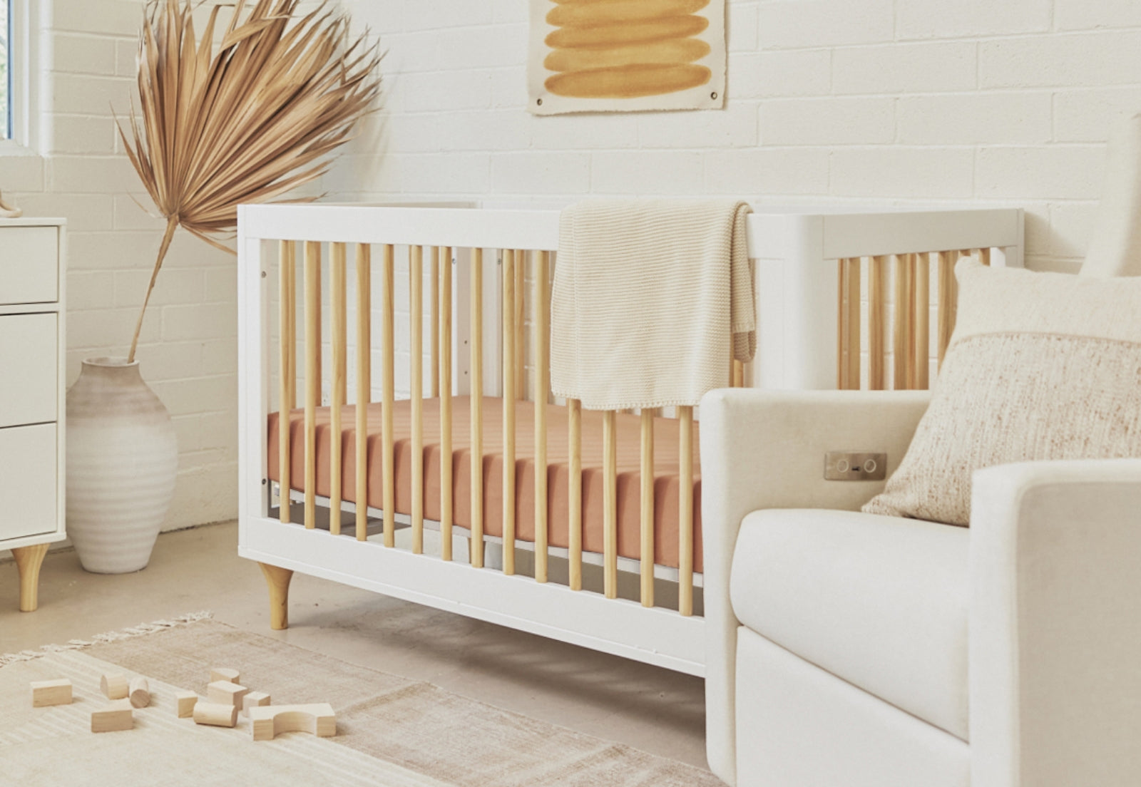 Cozy nursery room with a modern white crib draped with a beige blanket, a comfortable armchair, and soft blocks on the floor, adorned with natural decor elements.