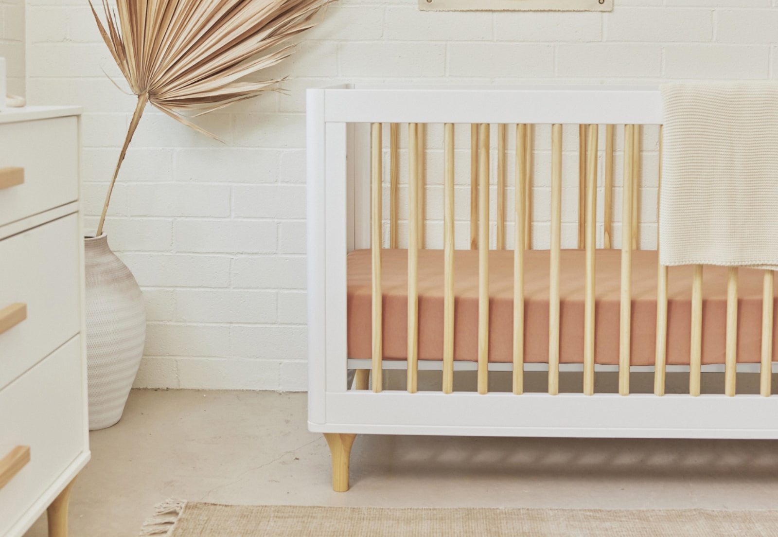 A modern nursery featuring a stylish white and wood crib against a white brick wall, complemented by a dried palm leaf decoration and minimalist furniture.