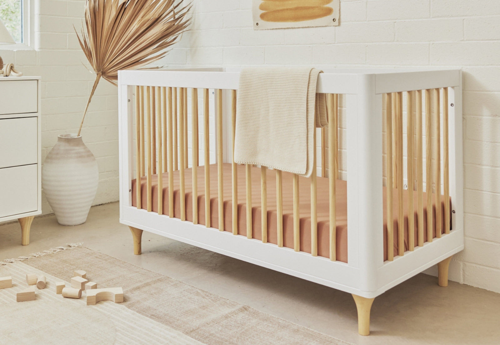 A modern wooden crib with a white frame and natural wood legs in a bright nursery, accessorized with a beige blanket, wooden toys on the floor, and a large vase with dried foliage. 999-8502-WHN
