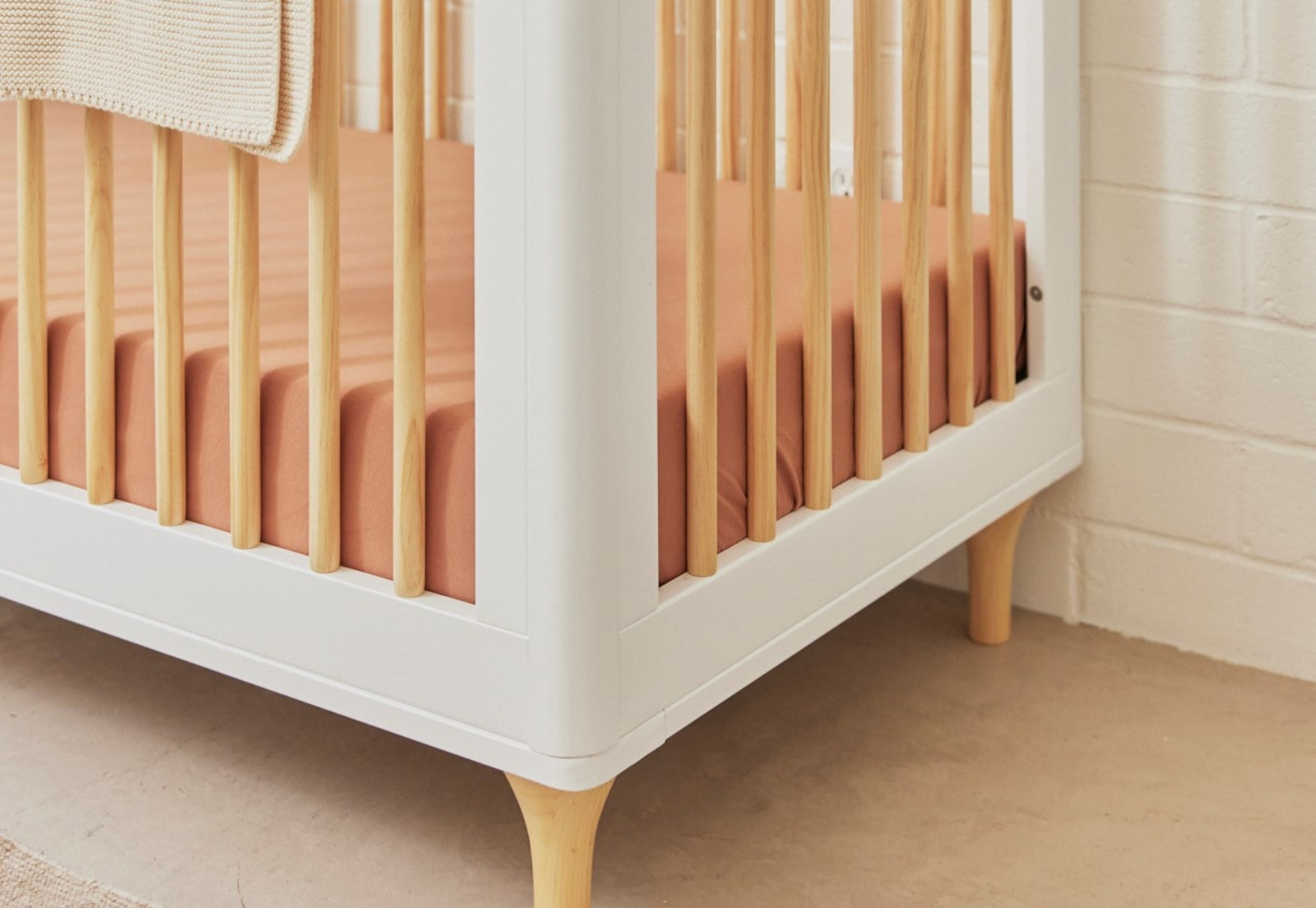Close-up of a stylish white baby crib with wooden legs and a peach-colored mattress, set against a neutral wall and floor.
