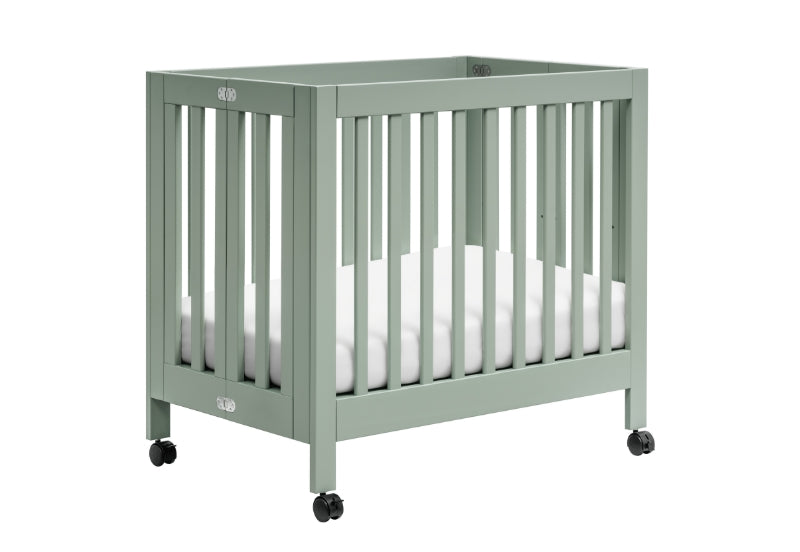 Sage green baby crib on wheels with white mattress, featuring visible brand logos on the crib ends.