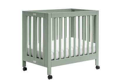 Sage green baby crib on wheels with white mattress, featuring visible brand logos on the crib ends.