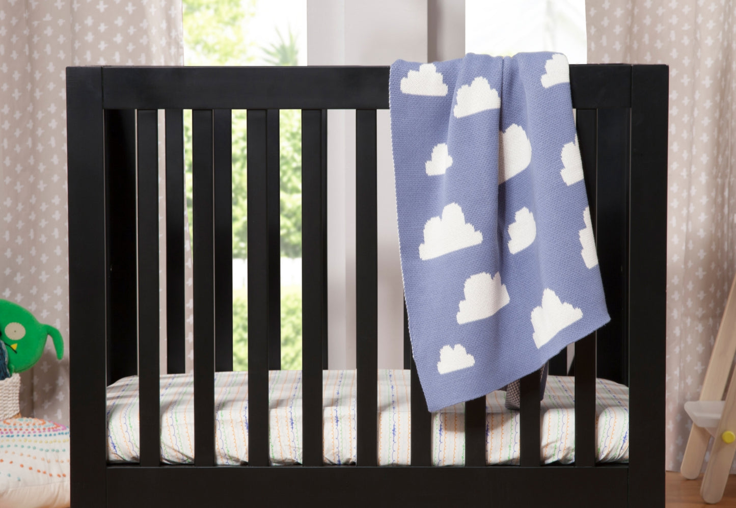 A cozy nursery featuring a black crib with a cloud-patterned blue blanket and colorful bedding, set by a window with sheer curtains.