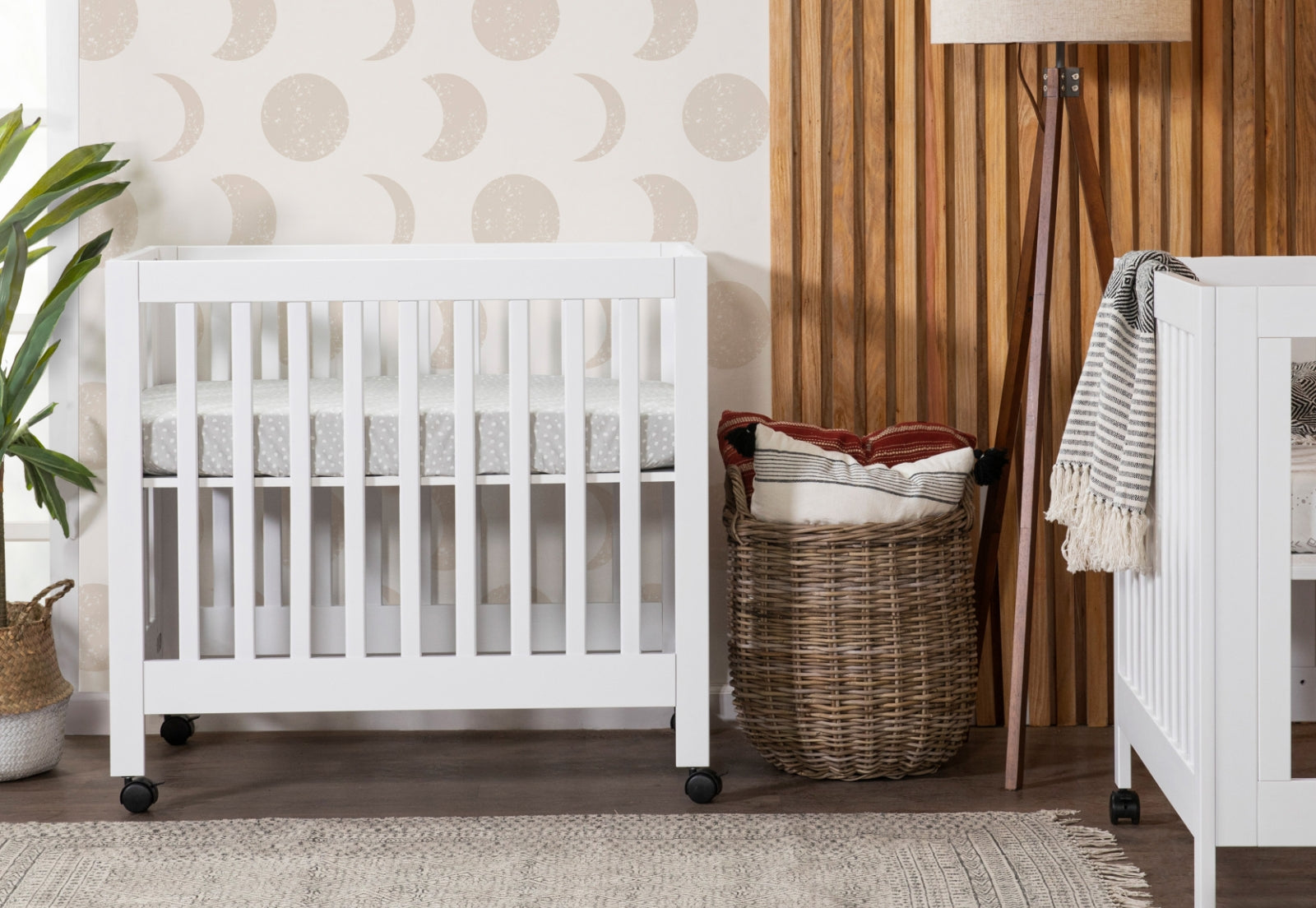 Stylish nursery featuring a white modern crib, decorative wicker basket with blankets, and wooden wall panels, set against a moon-patterned wallpaper. 999-8505-WHT