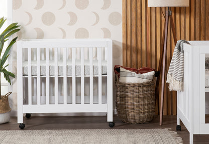 Stylish nursery featuring a white modern crib, decorative wicker basket with blankets, and wooden wall panels, set against a moon-patterned wallpaper. 999-8505-WHT