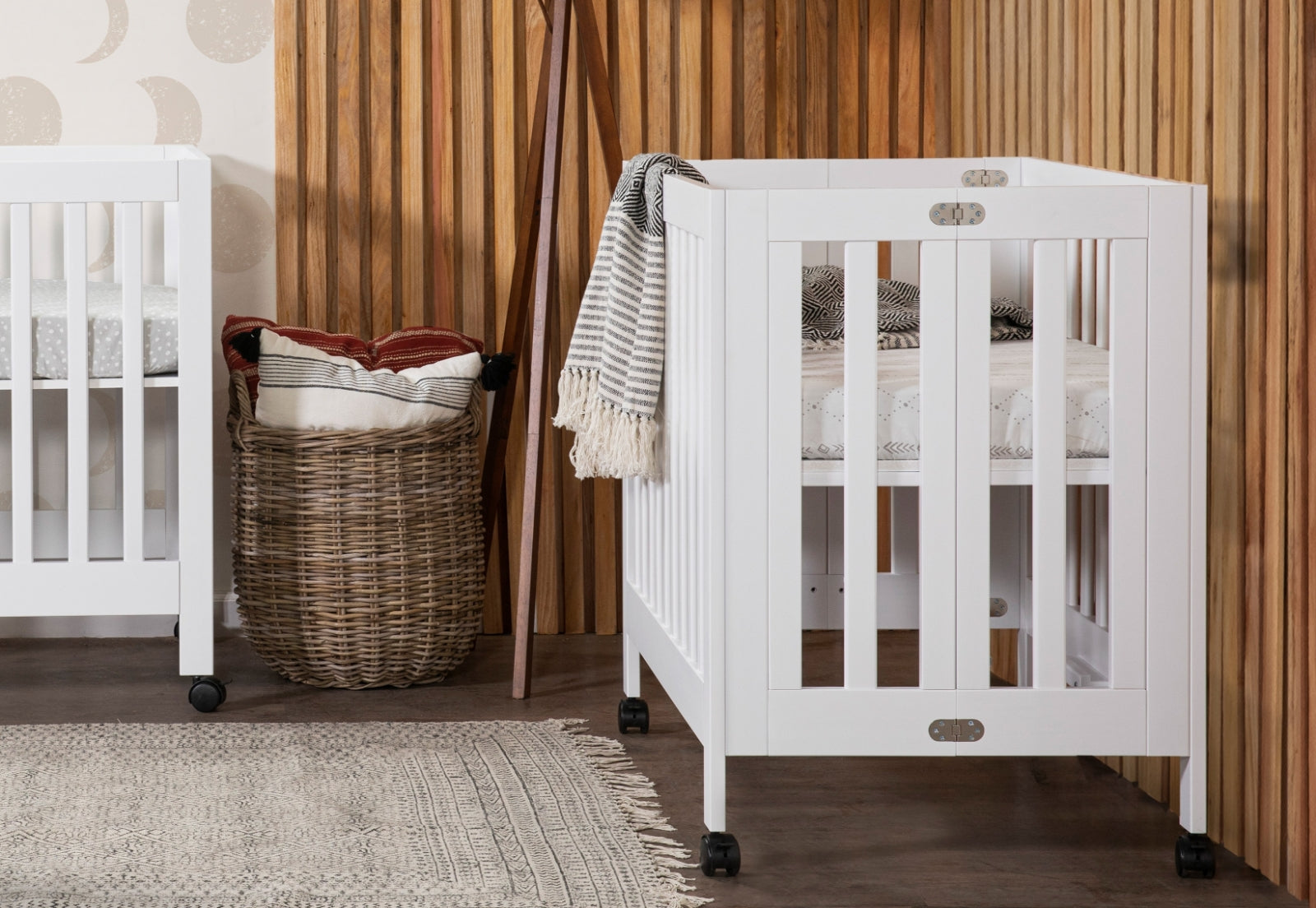 Two white baby cribs in a room with a wooden wall and a patterned blanket hanging off one crib, placed near a wicker basket with blankets.