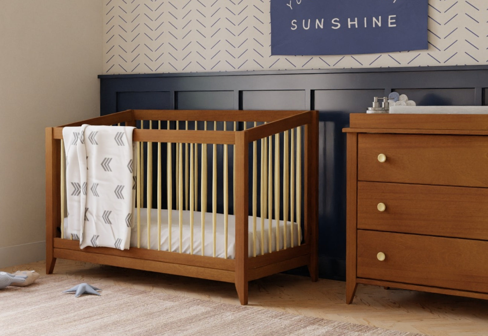 A well-organized nursery featuring a wooden crib with a patterned blanket, a matching dresser, and a wall decor saying "YOU ARE MY SUNSHINE" above a paneled wall. 999-8503-CHN