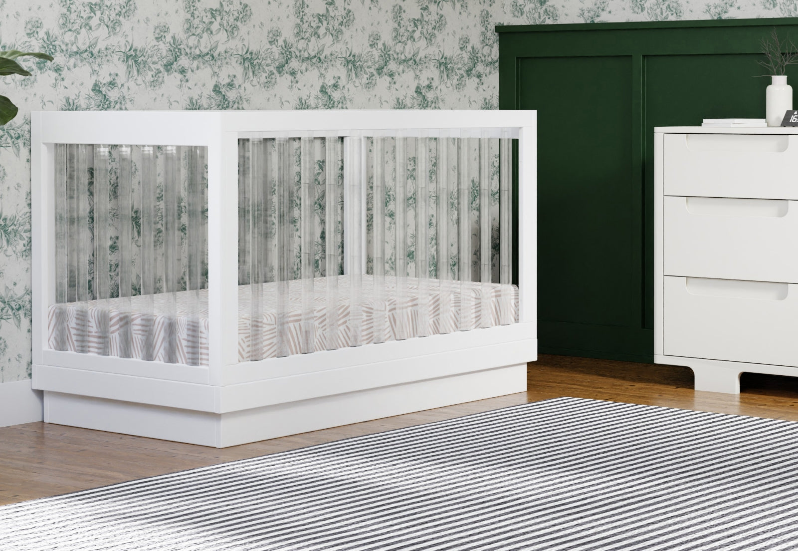 Modern white crib in a stylish nursery with floral wallpaper and dark green accents.