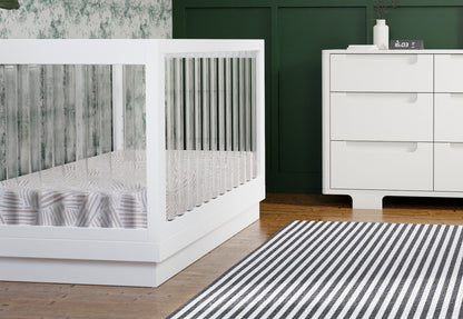 Modern nursery featuring a stylish white baby crib beside a sleek white dresser, complemented by a green wall and a striped rug.