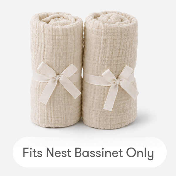 Two cream-colored, textured Newton Baby Nest Bassinet Extra Sheets are rolled and tied with matching fabric bows on a white background. Text below reads “Fits Nest Bassinet Only.” Soft cotton muslin ensures cozy comfort for your baby’s sleep.