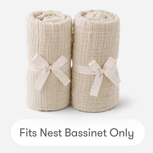 Two cream-colored, textured Newton Baby Nest Bassinet Extra Sheets are rolled and tied with matching fabric bows on a white background. Text below reads “Fits Nest Bassinet Only.” Soft cotton muslin ensures cozy comfort for your baby’s sleep.