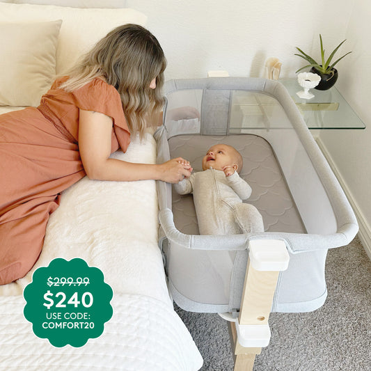 A woman in orange holds hands with a smiling baby lying in the Newton Baby Bassinet & Bedside Sleeper. Text: $240 (was $299.99) with code COMFORT20. A nightstand with a plant sits nearby.