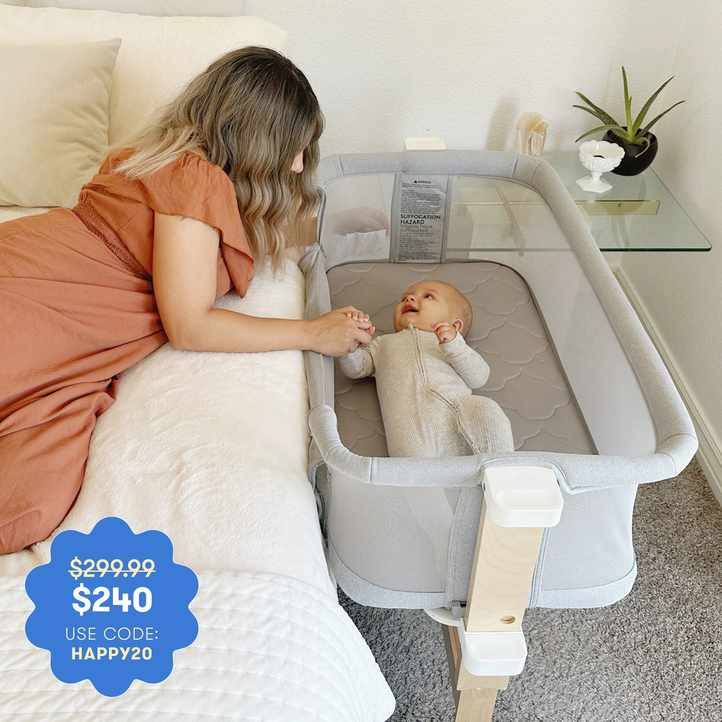 A woman in an orange dress lies on a bed, holding hands with a smiling baby in the Newton Baby Bassinet & Bedside Sleeper. Price: $299.99 $240 with code HAPPY20.