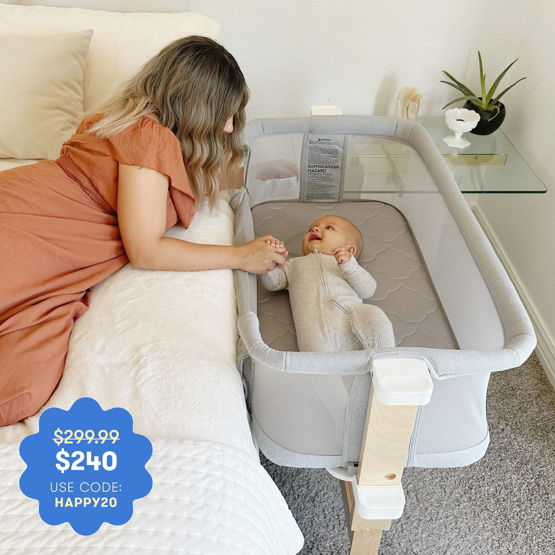 A woman in an orange dress lies on a bed, holding hands with a smiling baby in the Newton Baby Bassinet & Bedside Sleeper. Price: $299.99 $240 with code HAPPY20.