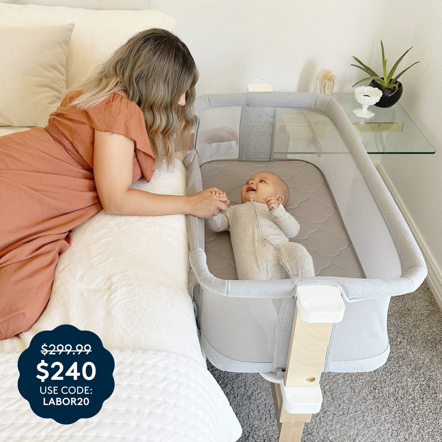 A woman wearing a brown dress lies on a bed, holding hands with a smiling baby resting in the Newton Baby Bassinet & Bedside Sleeper in gray. The price tag displays $299.99 crossed out, now $240 with code LABOR20. A glass shelf decorated with decor is visible in the background.